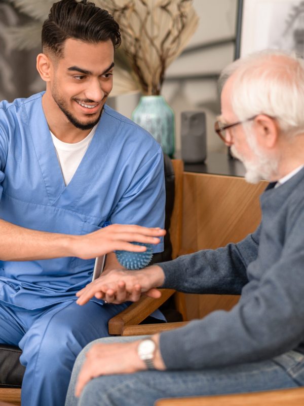 Smiling friendly physiatrist looking at an aged patient while massaging the back of his hand